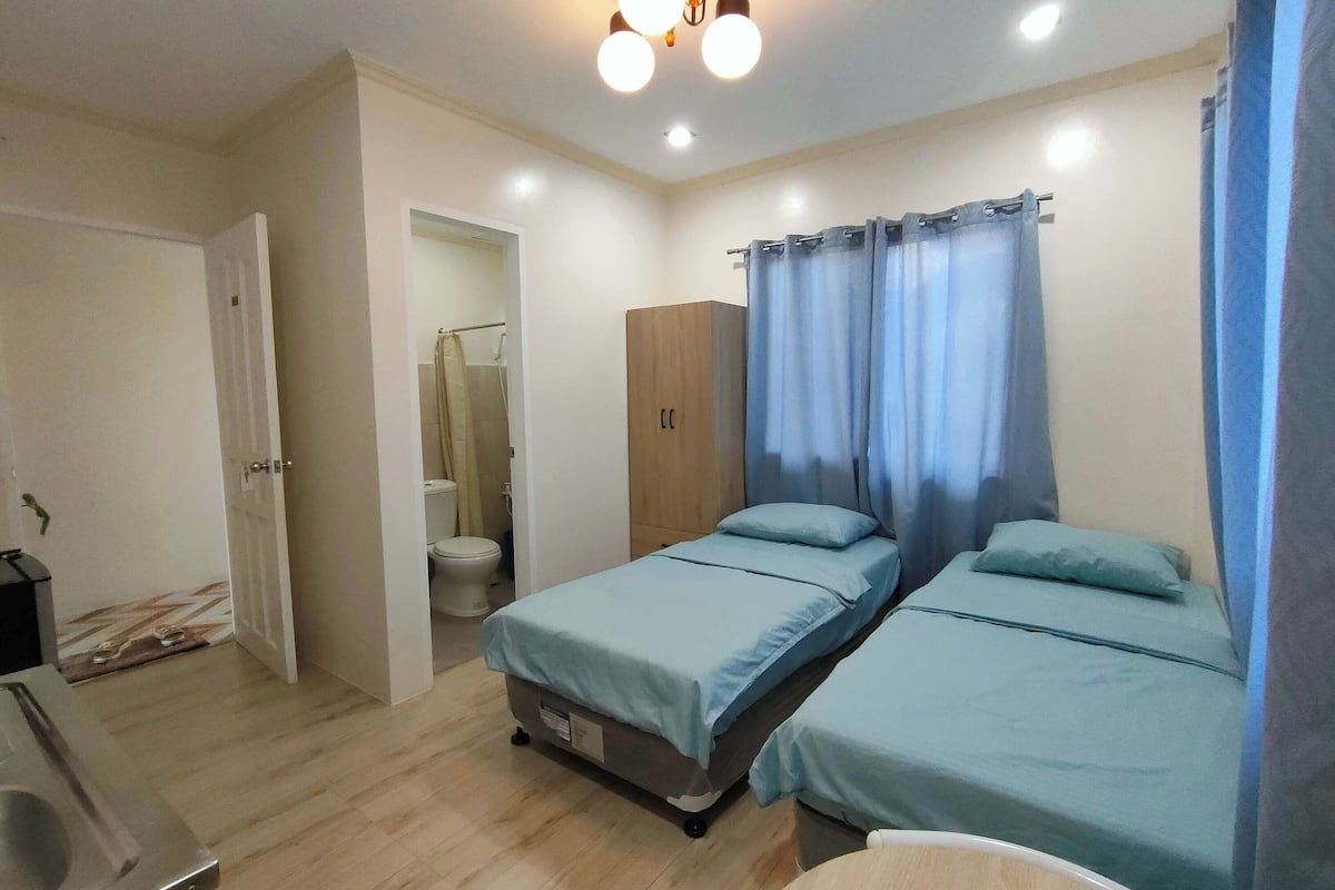 A small, well-lit bedroom with two single beds, a wooden wardrobe, and a bathroom visible through an open door.