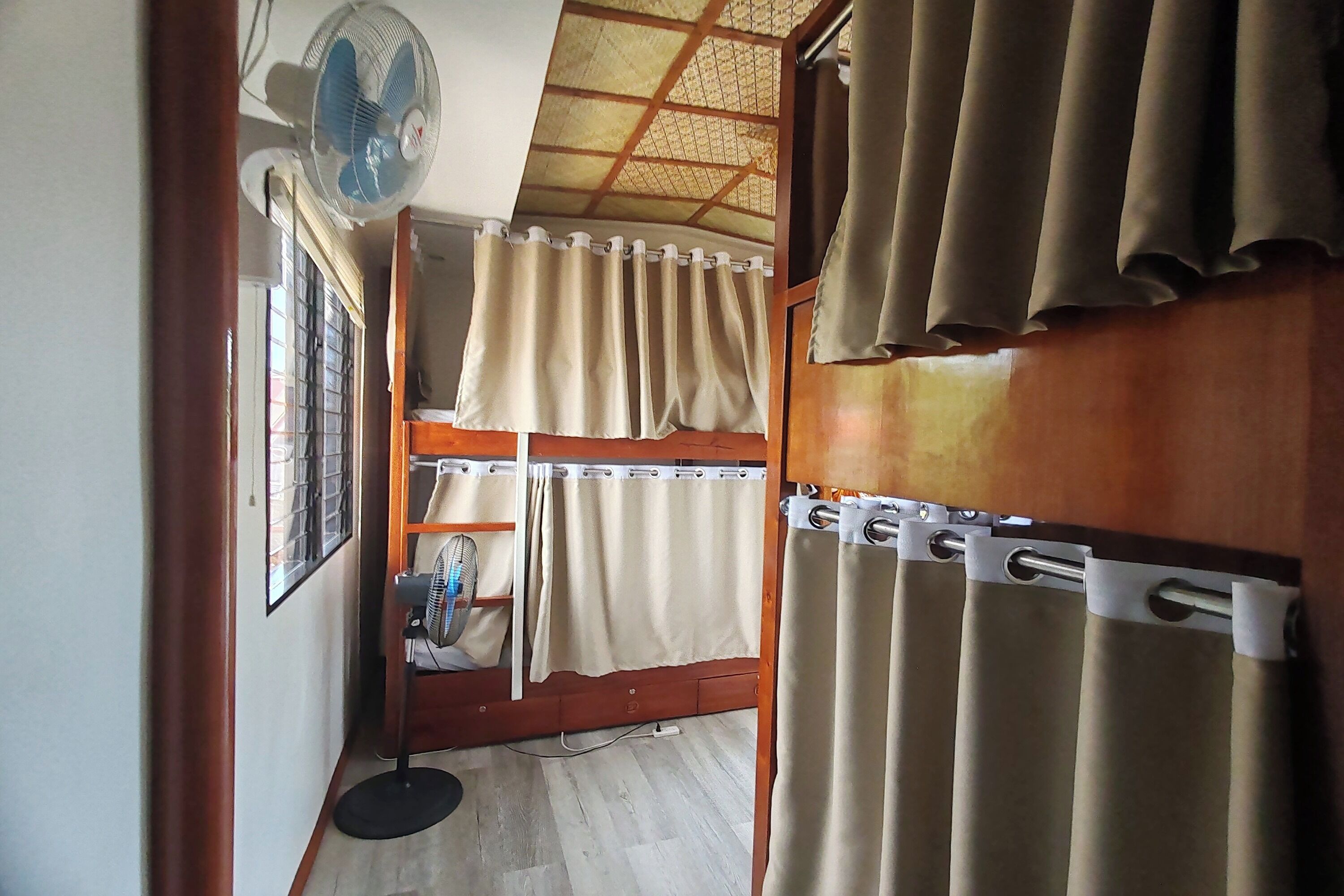 A room with bunk beds, a standing fan, a window with bars, and curtains.