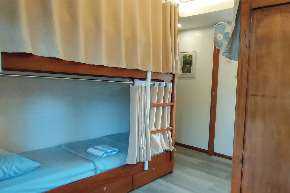 A bunk bed with a blue mattress and beige curtains, a wooden wardrobe, and a wall-mounted fan.
