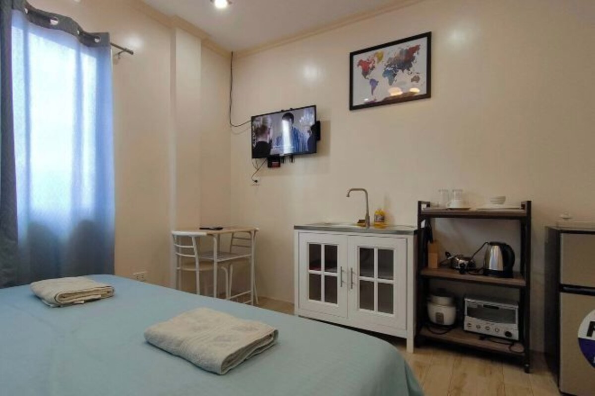Une chambre d’hôtel avec un lit, une petite table, un meuble contenant des appareils de cuisine et un téléviseur fixé au mur.