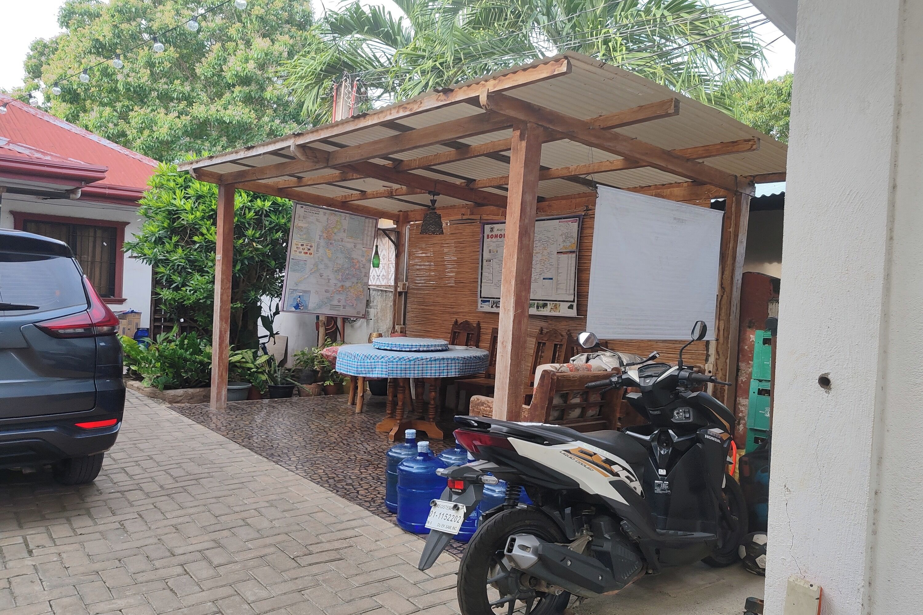 A covered outdoor area with a table and chairs, a motorcycle, and large blue water containers.