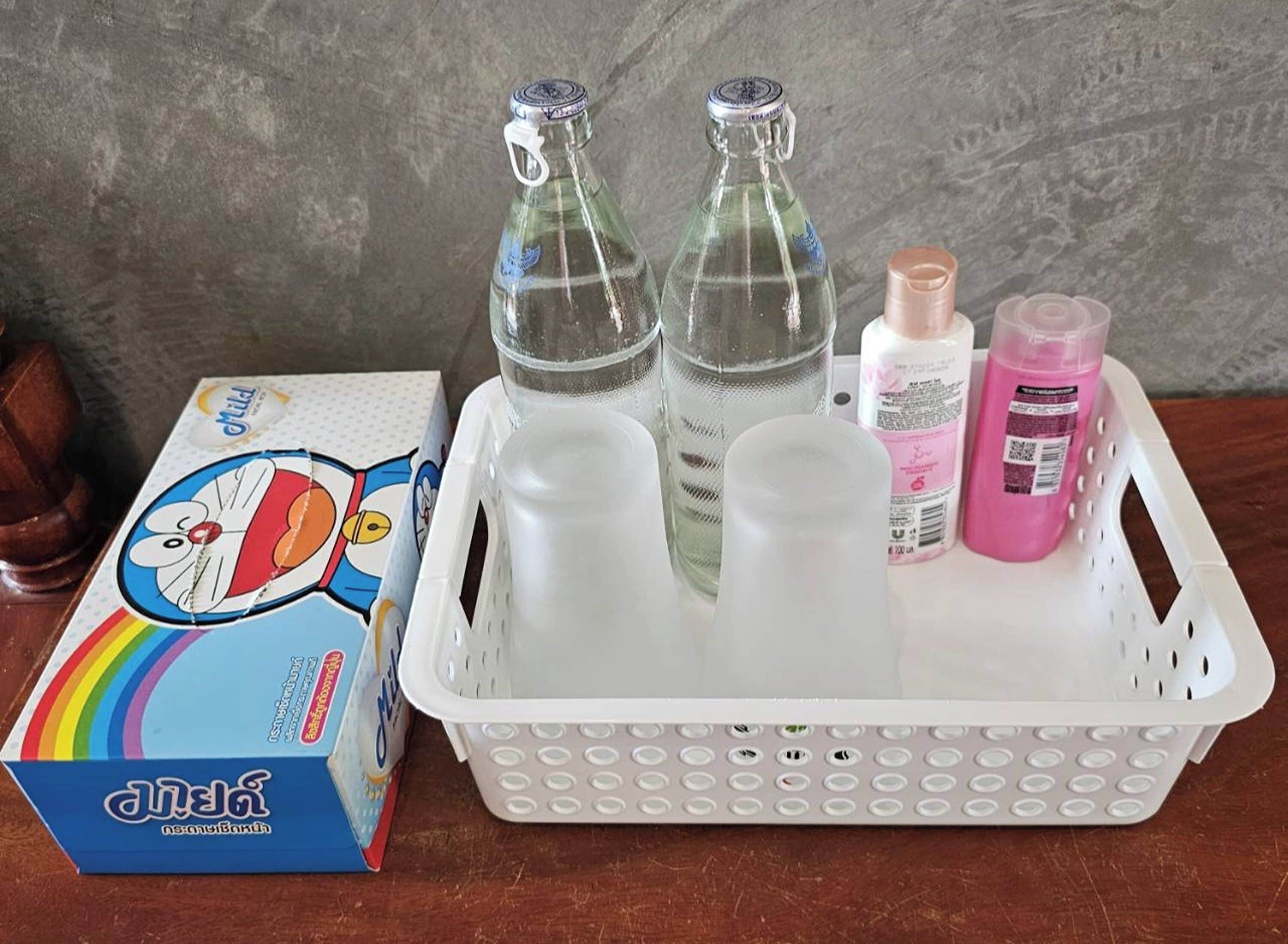 A plastic basket containing two glass bottles, a plastic cup, and two bottles of pink liquid on a wooden surface with a cartoon box.