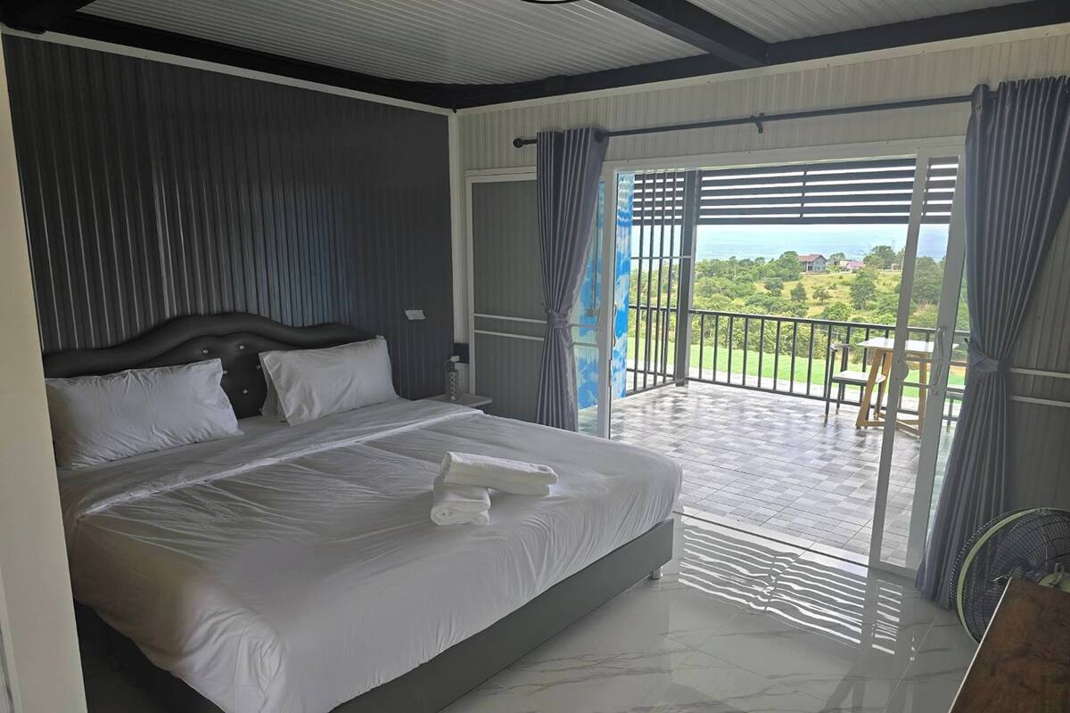 A spacious bedroom with a large bed, a view of the outdoors, and a balcony with a railing.