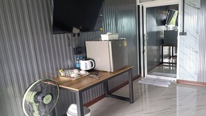Een compacte kamer met een houten tafel, een waterkoker, een kleine koelkast, een ventilator en een schuifdeur naar een balkon.