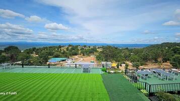 A tennis court with a green surface, surrounded by a fence, and a view of a hilly landscape with trees and buildings in the distance.