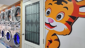 A row of industrial-style laundry machines with numbered slots, a glass door with a geometric pattern, and a cartoon tiger mural on the wall. - Reezq Hostel (Bangkok)