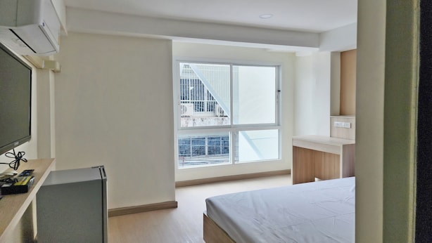 A bedroom with a bed, a desk, a television, and a window with a view of a building. - Reezq Hostel (Bangkok)