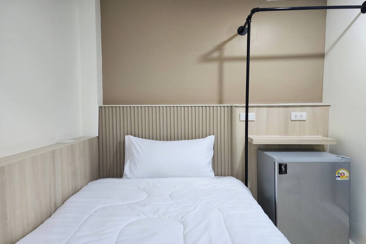 A small, neatly made hotel room with a single bed, a compact refrigerator, and a simple wall-mounted shelf.