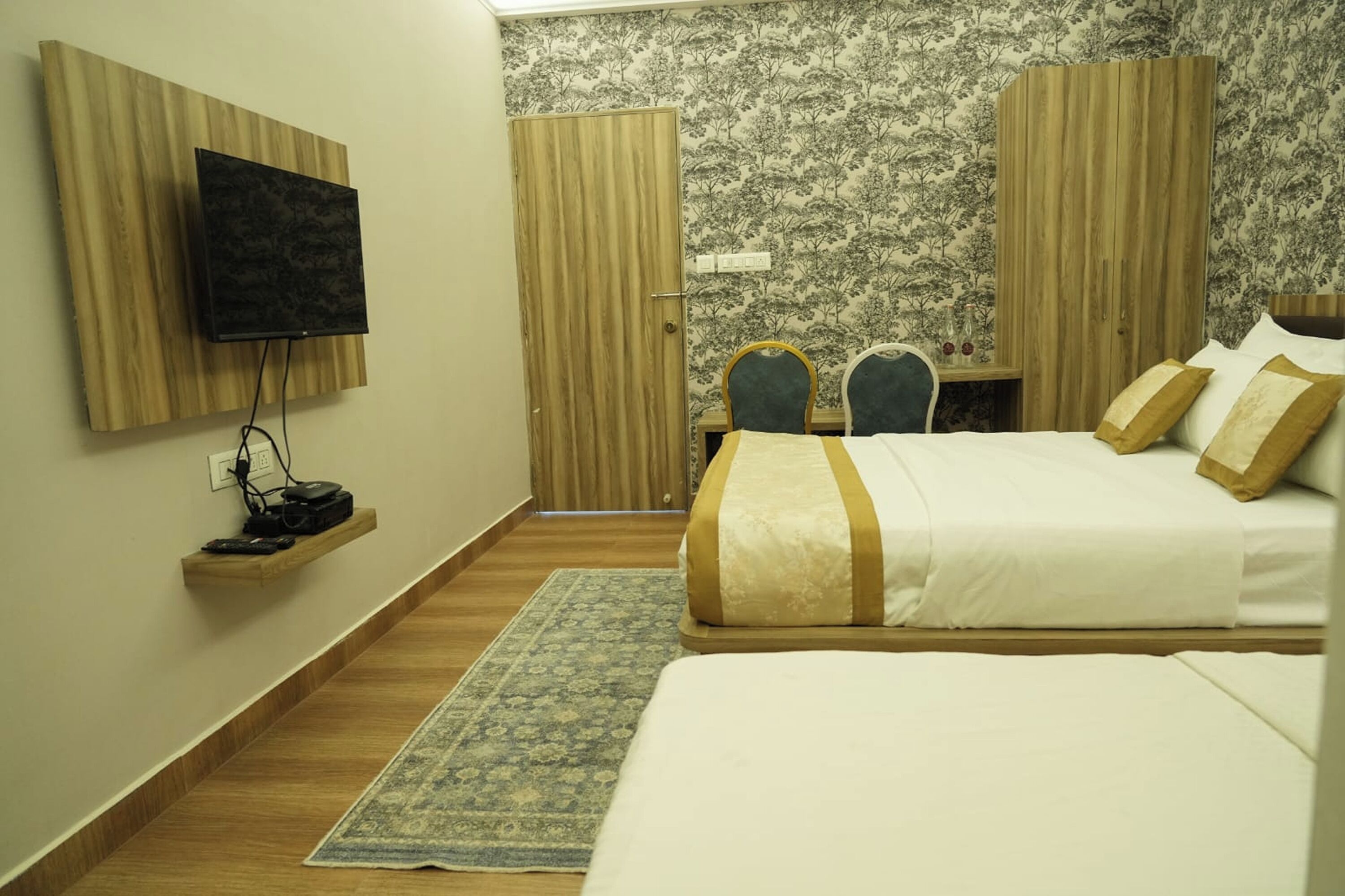 A bedroom with a bed, a TV mounted on a wooden panel, a desk with two chairs, and a wardrobe.
