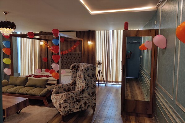 A room with a bed, a floral armchair, a wooden floor, a mirror, and a window with curtains. - Vilasini Inn (Madduru)