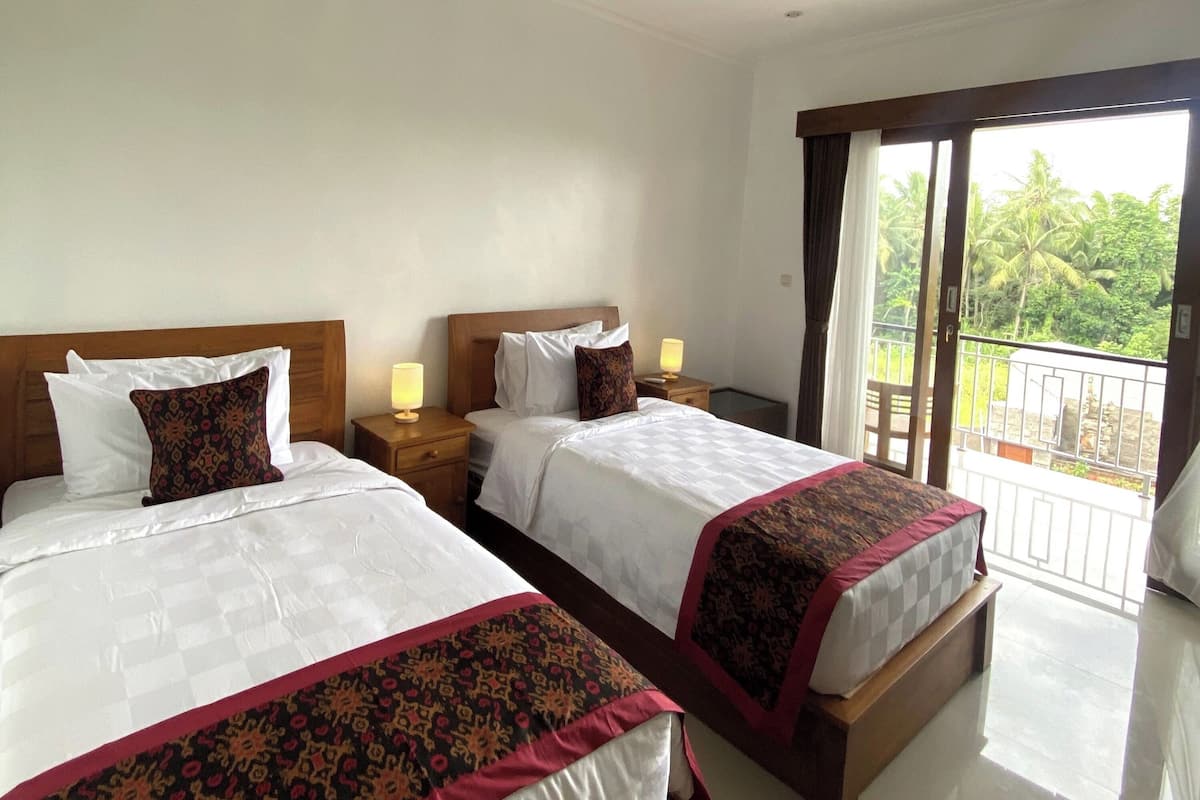 A hotel room with two beds, each with a wooden headboard, white linens, and decorative pillows. There are bedside tables with lamps, and a balcony with a view of greenery.