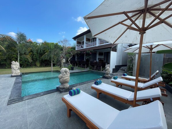 A poolside area with lounge chairs and umbrellas, a statue, and a building with a balcony in the background. - Nivaluma By Zuzu (Mas)