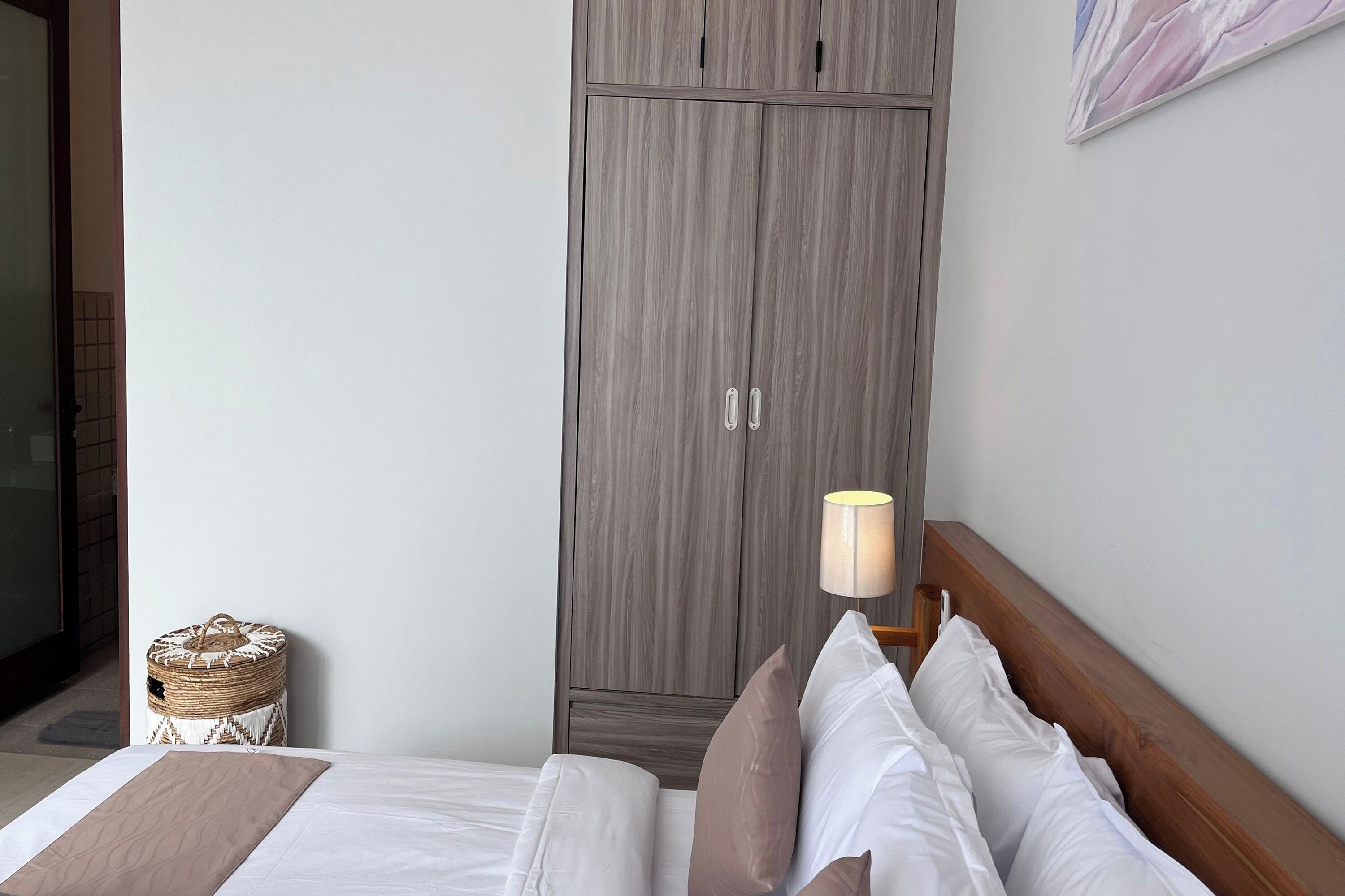 Ein Schlafzimmer mit Bett, Kissen, einer Nachttischlampe, einem Kleiderschrank und einem kleinen, runden Hocker.