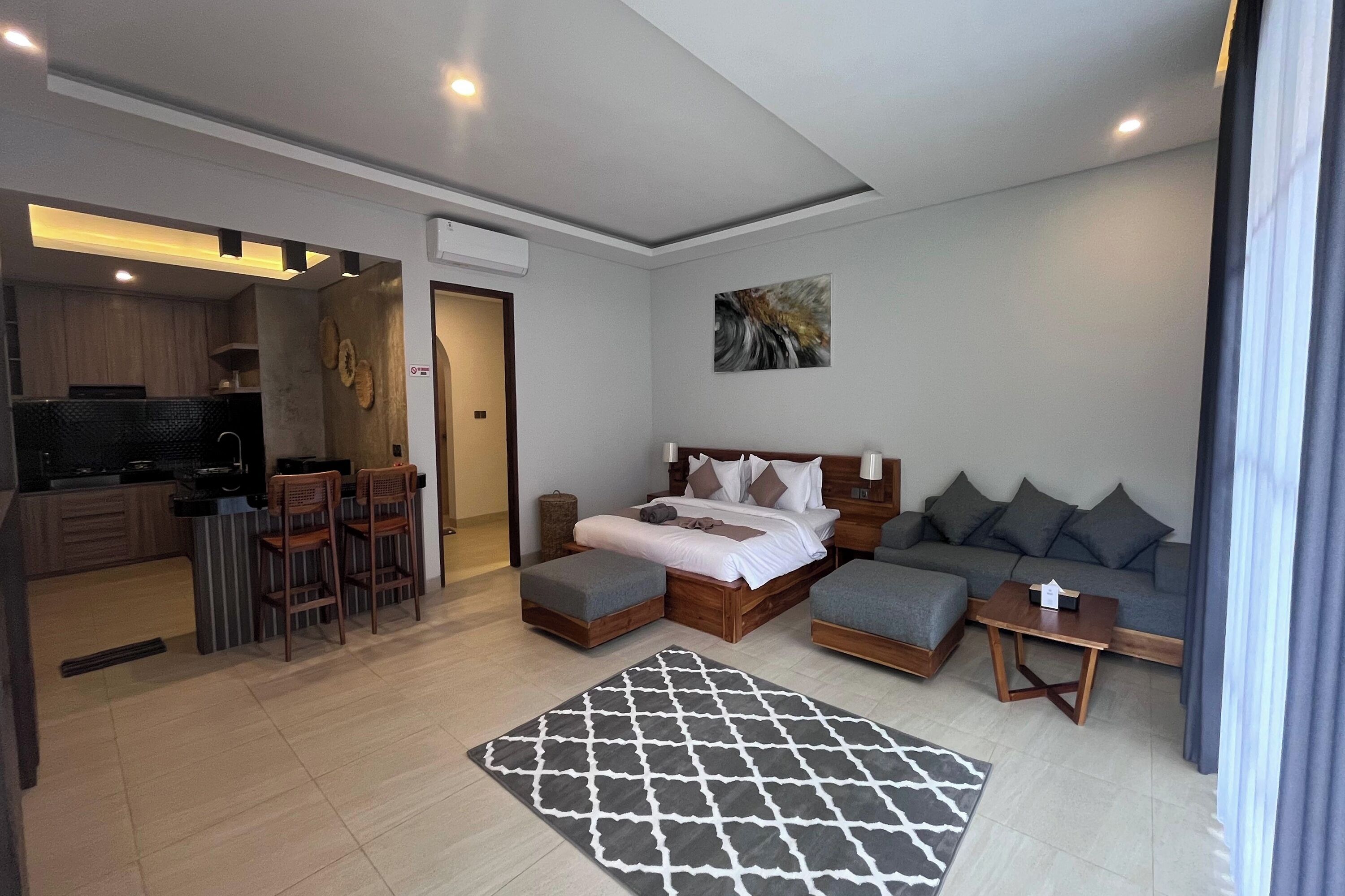 A modern hotel room with a bed, sofa, ottoman, and a small table. There is a kitchen area with a counter and chairs.
