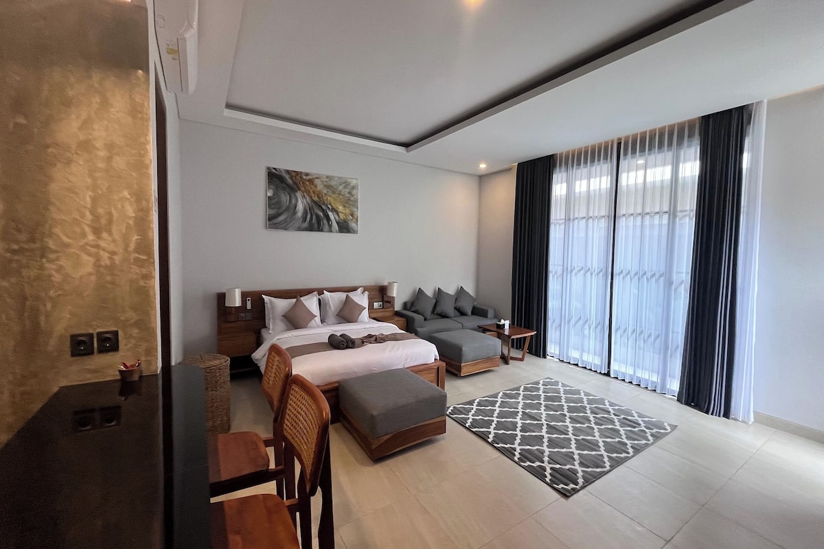 A modern hotel room with a large bed, a sofa, a desk, and a view of the outdoors.