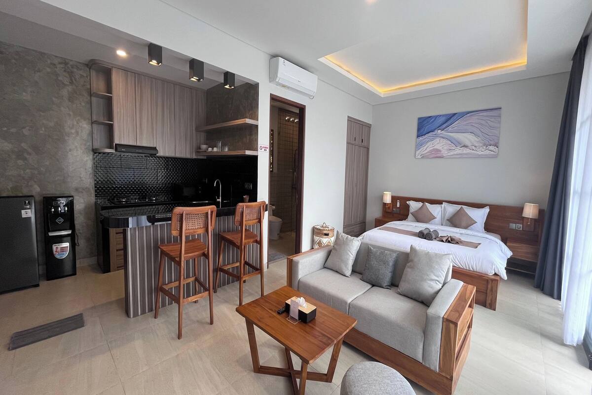 A modern hotel room with a kitchenette, a sofa bed, a wooden coffee table, and a painting on the wall.