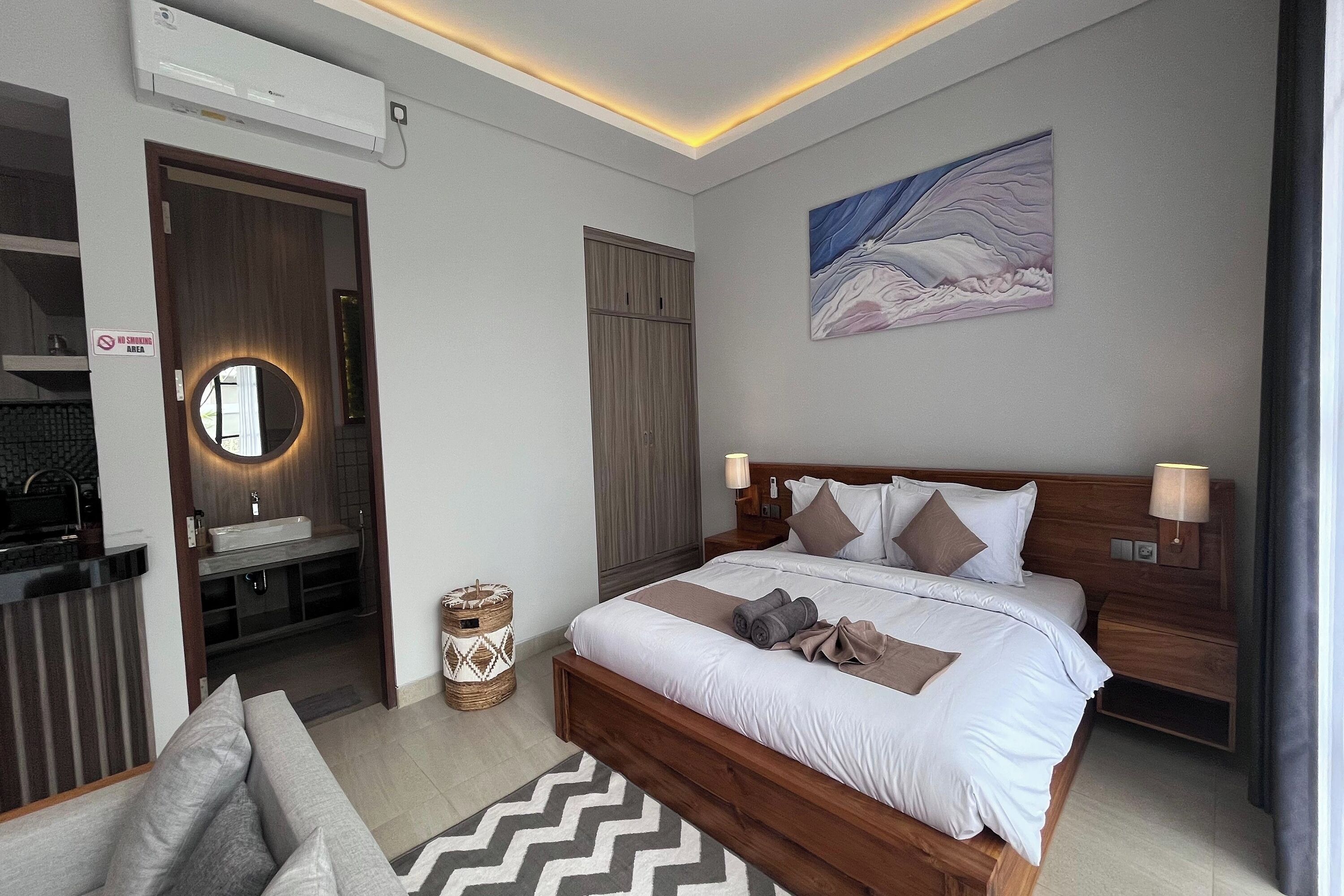A modern hotel room with a wooden bed, a round mirror, a painting on the wall, and a separate bathroom area.