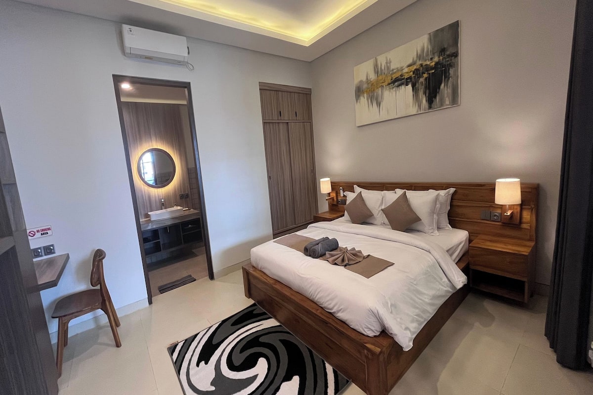 A modern hotel room with a wooden bed, bedside lamps, a round mirror, a painting, and a small desk with a chair.