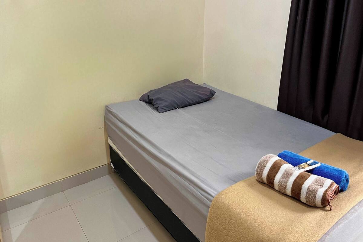 A small, neatly made bed with a grey sheet and a single pillow, a rolled towel on the bed, and a beige blanket on the floor.