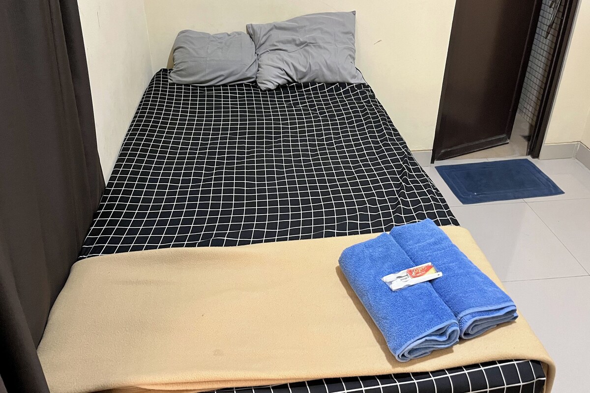 A single bed with a black and white checkered bedspread, two pillows, a beige blanket, and a blue towel on a blue mat.