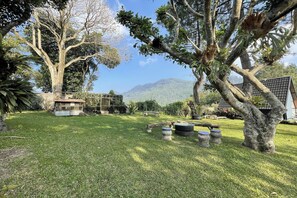 A well-maintained garden with a large tree, a small structure, and a clear view of a mountain in the background. - Villa 325 By Zuzu (Lembang)