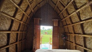 A room with a thatched roof, a bed with white linens, and a view of a garden through an open door.