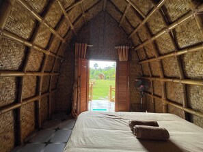A room with a thatched roof, a bed with white linens, and a view of a garden through an open door.