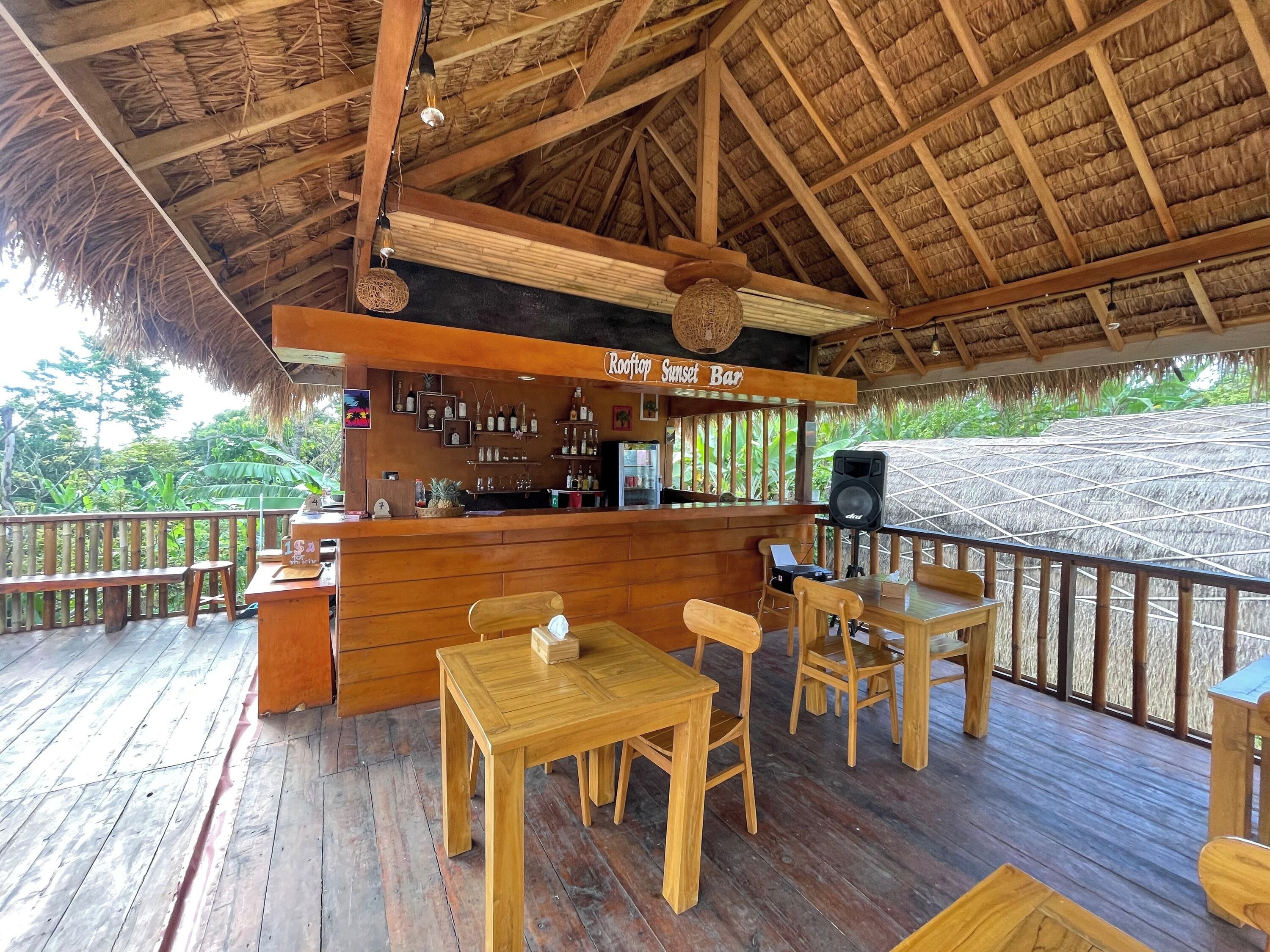 Een houten terras met rieten dak, een bar en tafels en stoelen. Op het bord staat 'Roof Top Sunset Bar'.