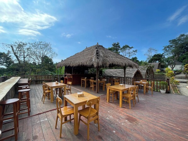 An outdoor dining area with wooden tables and chairs, a thatched-roof structure, and a view of the surrounding greenery.