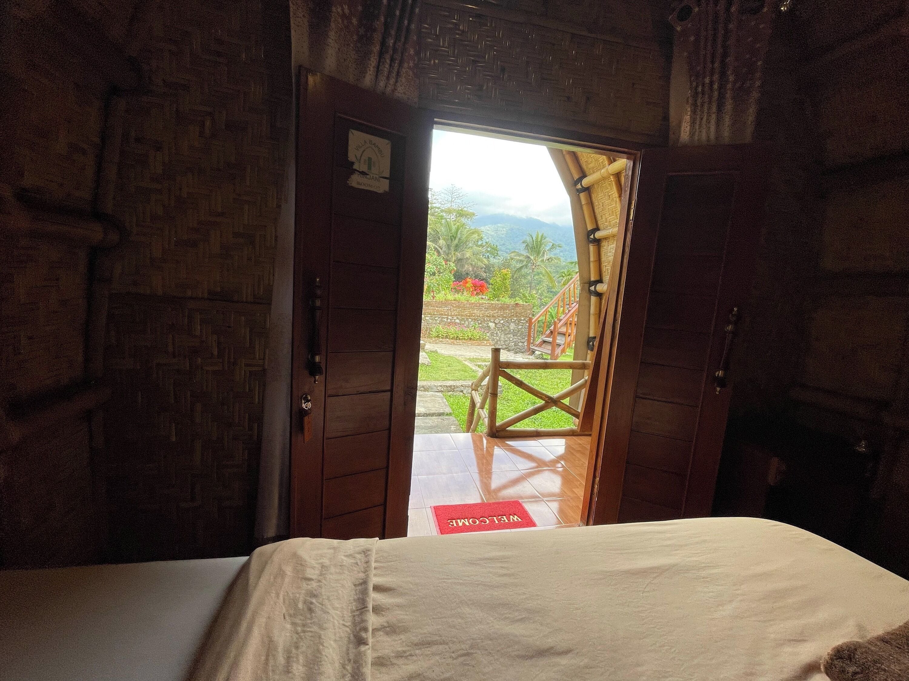 A room with a bed, a wooden door leading outside, and a red mat with "WELCOME" on it.
