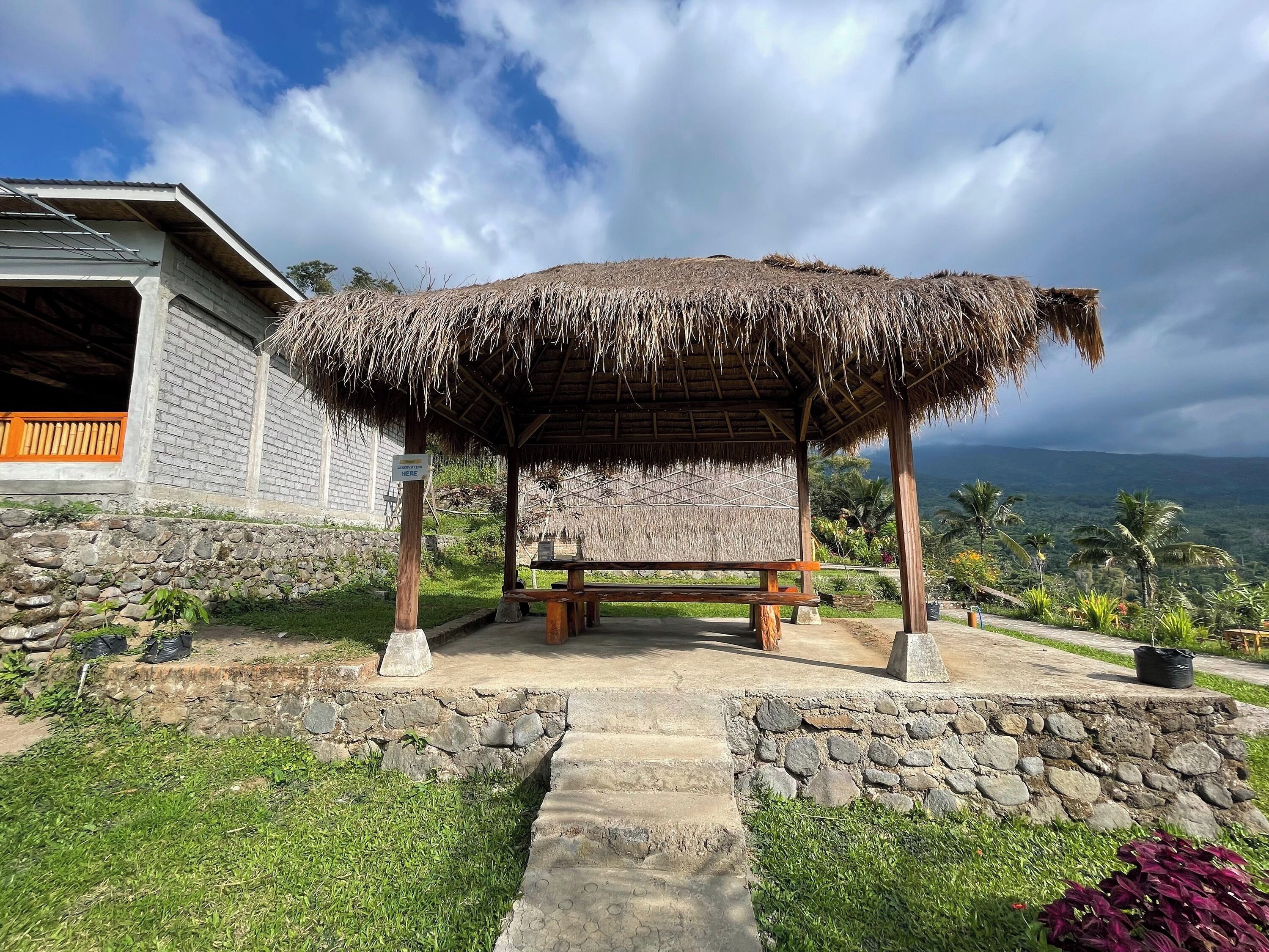 A thatched-roof gazebo with a bench, surrounded by stone walls and greenery, with a view of a mountain range.