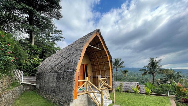 A thatched-roof hut with a wooden staircase leading to a door, surrounded by lush greenery and a stone wall.