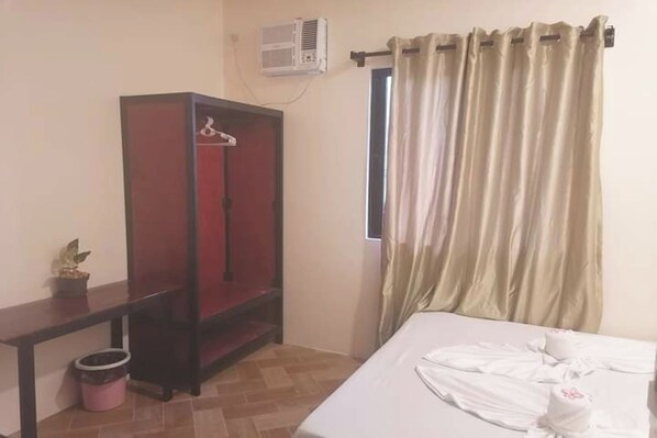 Una habitación con cama, un armario, una mesita con una planta, un cubo de basura y una ventana con cortinas.