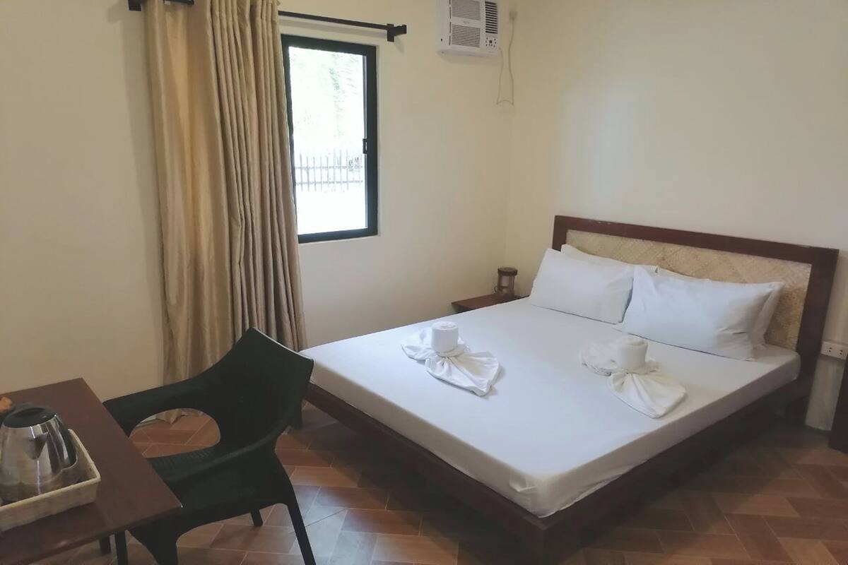 Ein Hotelzimmer mit Bett, Stuhl, Tisch mit Wasserkocher und Fenster mit Vorhängen.