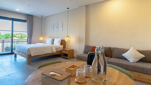 A modern hotel room with a large bed, a sofa, a dining table, and a balcony with a view of greenery. - Kelaya Guesthouse (Denpasar)