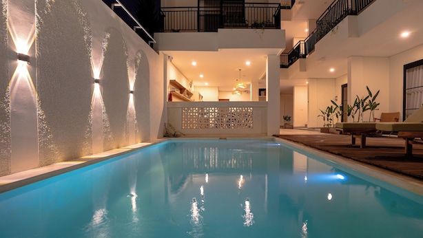 A swimming pool with a wooden deck and lounge chairs, a balcony with a railing, and a kitchen area visible through a partition. - Kelaya Guesthouse (Denpasar)
