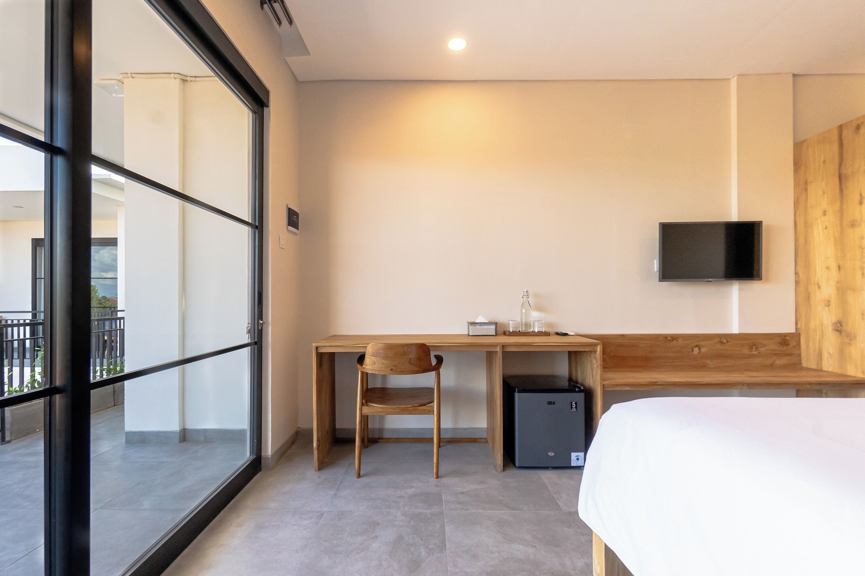 Een moderne hotelkamer met een houten bureau, een stoel, een bed, een aan de muur bevestigde televisie en een kleine, zwarte koelkast.