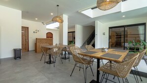 A modern dining area with wicker chairs and a wooden table, a staircase, and a large window. - Kelaya Guesthouse (Denpasar)