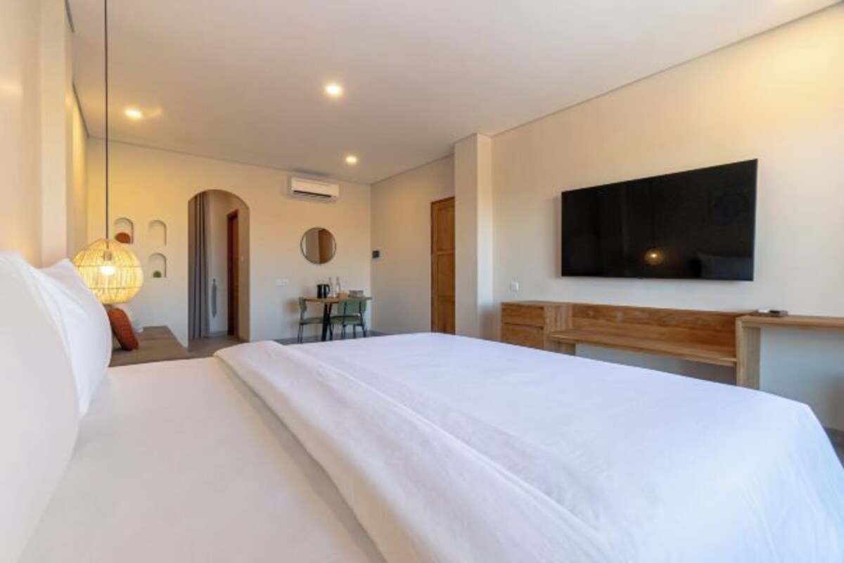 Quarto de hotel com uma cama grande, uma escrivaninha com cadeira, uma televisão na parede e uma mesinha de cabeceira com abajur.