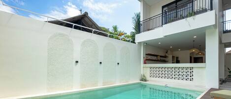 Una piscina con bordo in legno e lettini prendisole, un muro esterno bianco con pannelli decorativi e un balcone con ringhiera.