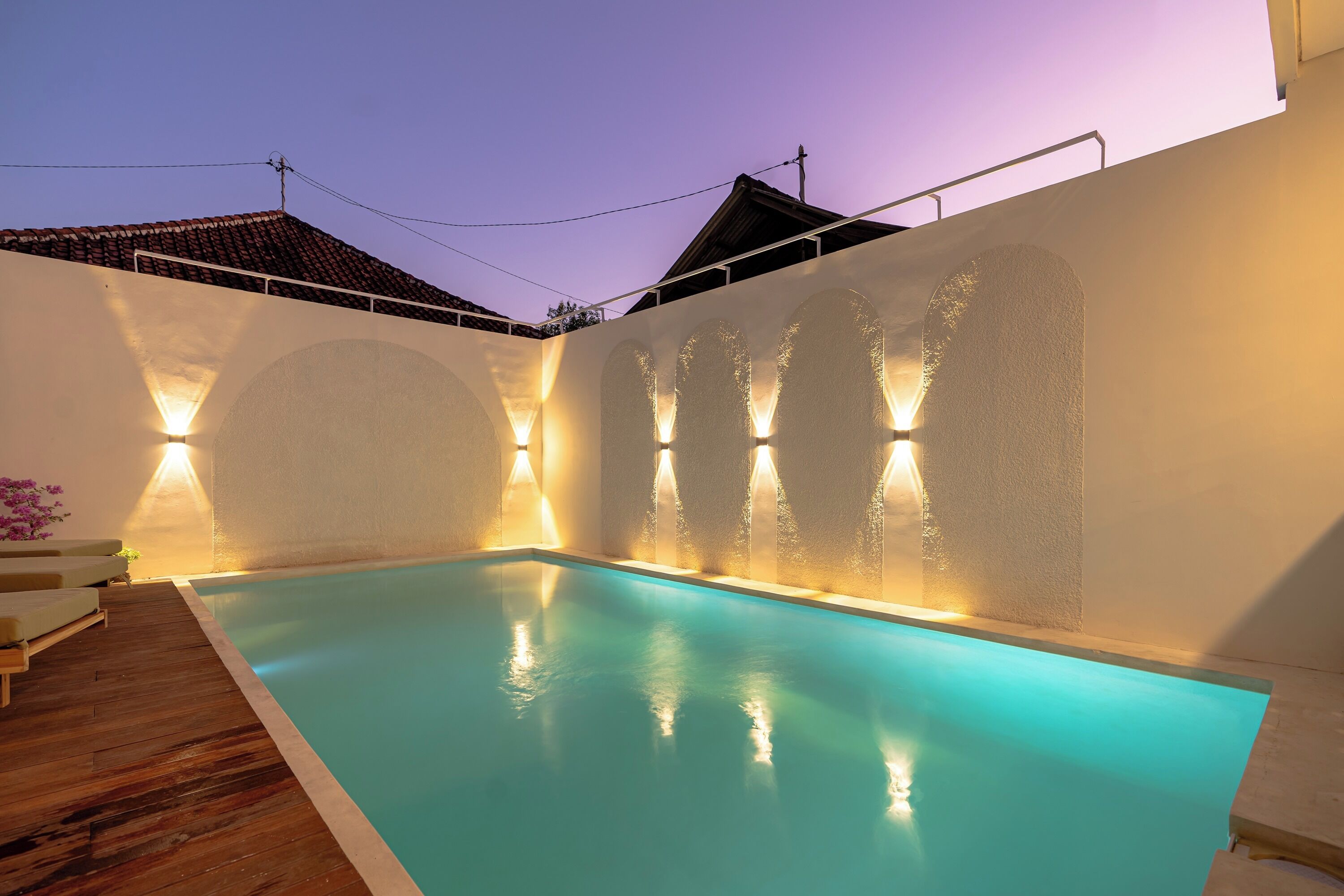 A swimming pool with a wooden deck and a white wall with illuminated arches.