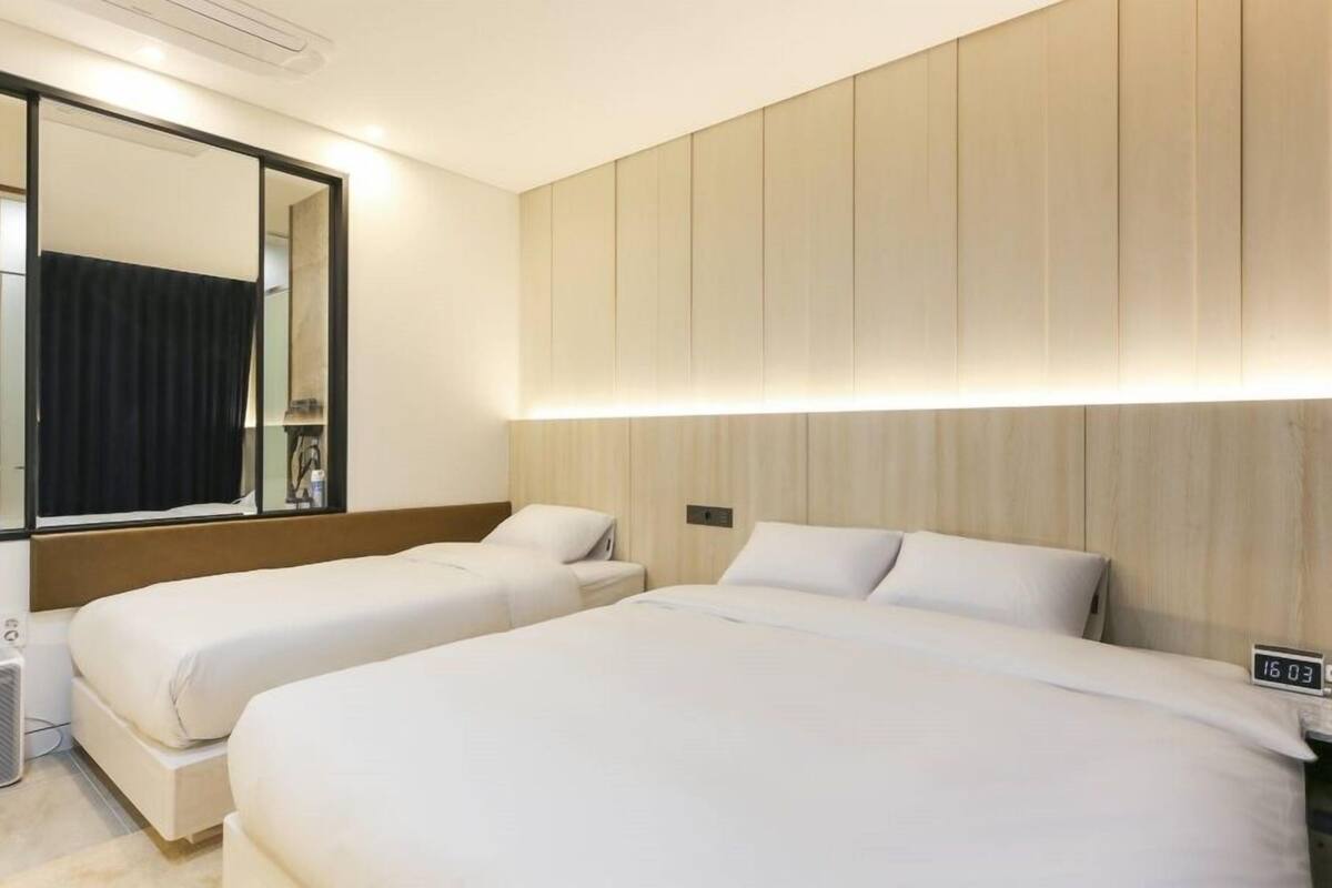 A hotel room with two beds, a large mirror, a nightstand with a digital clock, and a wall-mounted heater.