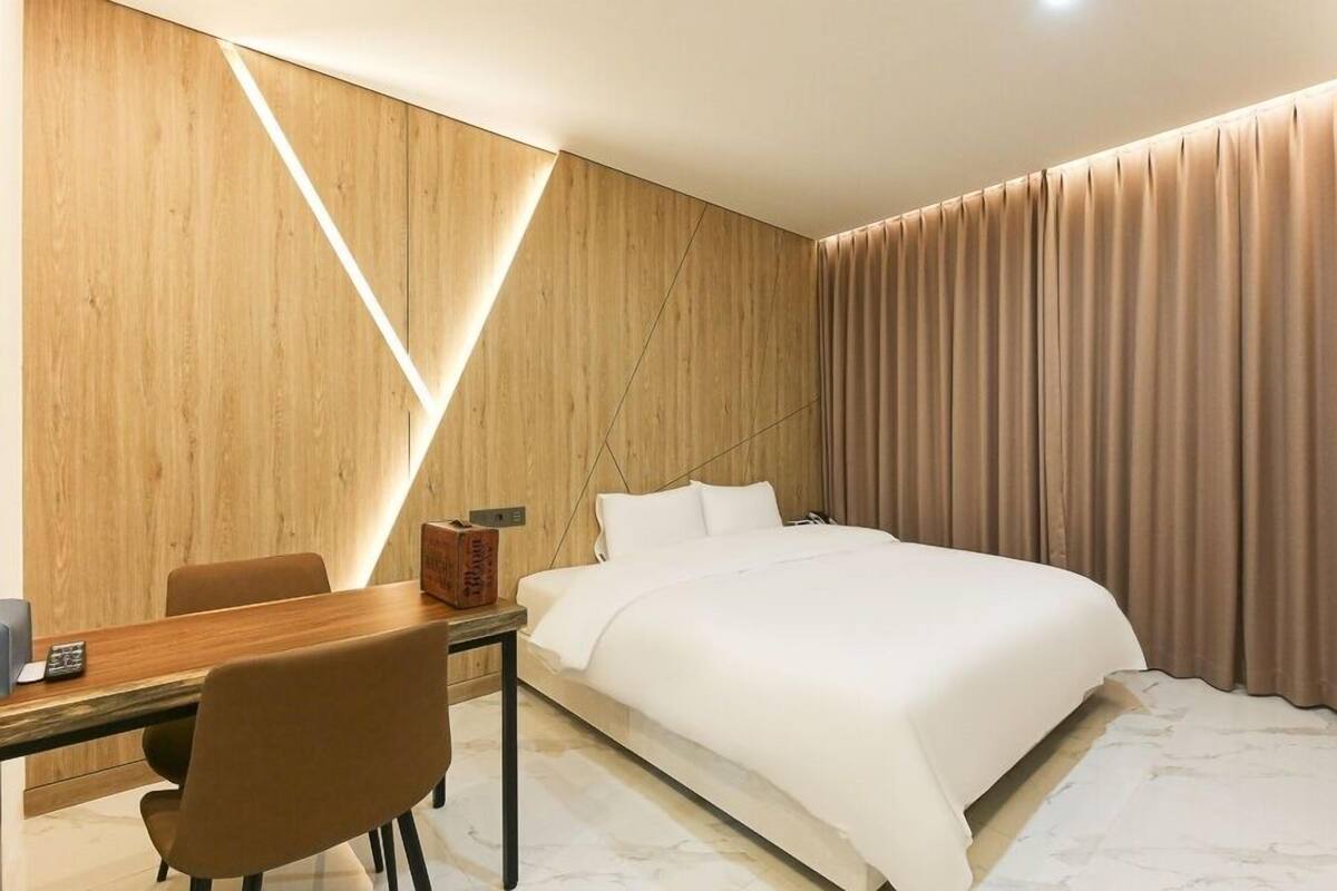 A modern hotel room with a wooden accent wall, a bed with white bedding, a desk with a chair, and a large window with curtains.
