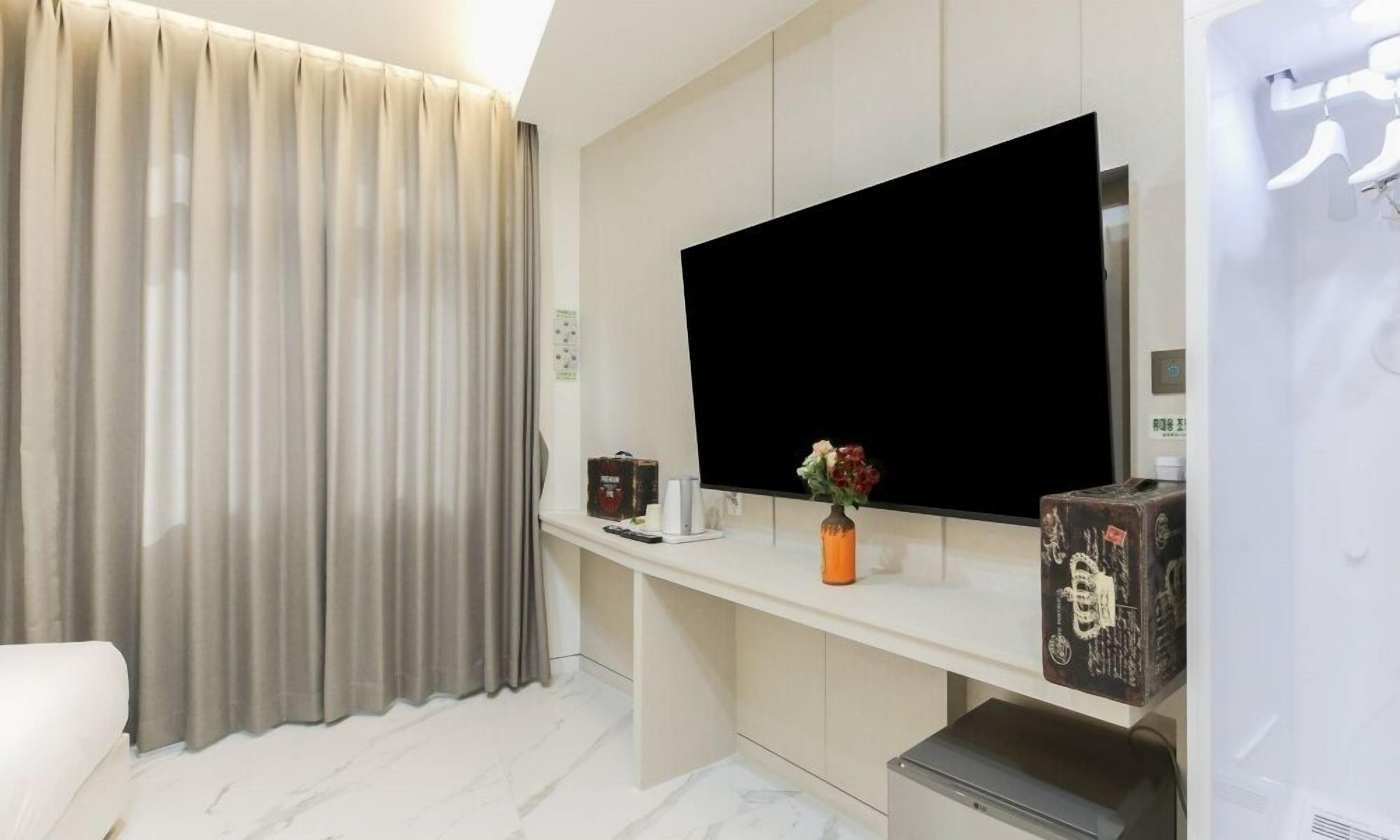 A modern hotel room with a flat-screen TV mounted on the wall, a small table with a vase of flowers, and a large window with curtains.
