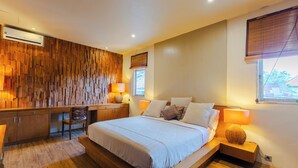A modern bedroom with a wooden wall panel, a bed with white linens, a desk, and a chair. - Villa Bella Vista by Pertama Stay (Seminyak)
