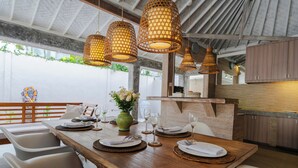 A dining area with a wooden table set for four, surrounded by white chairs with blue cushions. There are wicker pendant lights above the table, and a vase with flowers on the table. - Villa Bella Vista by Pertama Stay (Seminyak)
