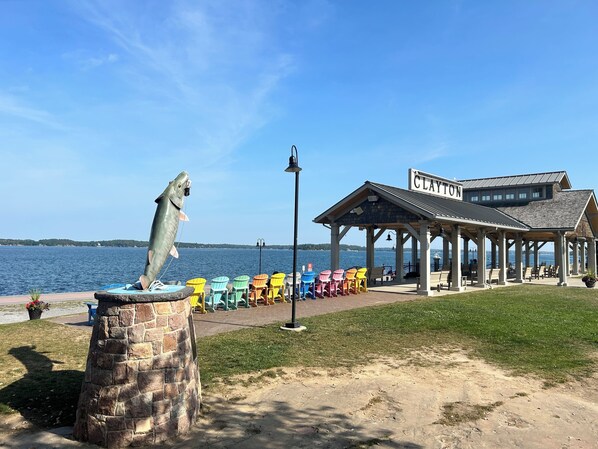 Une statue représentant un pêcheur, posée sur un piédestal en pierre au bord de l’eau, des chaises Adirondack colorées et un pavillon couvert appelé « Clayton ».