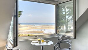 Una habitación con un amplio ventanal con vista a la playa, una mesita con tazas y un plato, y dos sillas.