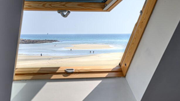 Vista desde una habitación con tragaluz, con vistas a una playa donde se ven personas y un velero en la distancia.
