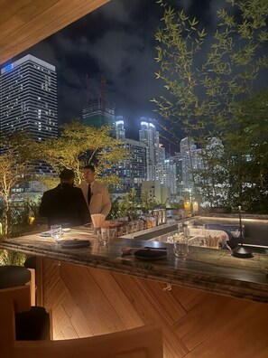 A bar area with a city view at night, featuring modern buildings and a wooden bar counter.